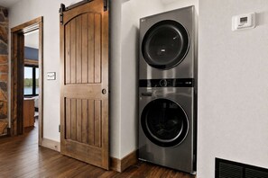 Un lavadero con entrada de doble puerta, lavadora y secadora de acero inoxidable, y vista a un dormitorio por una puerta entreabierta.