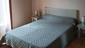 Um quarto com cama, mesas de cabeceira, luminária, quadro na parede e janela com cortinas.
