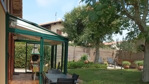 Um pátio com mesa e cadeiras, área coberta e jardim com gramado e plantas em vasos.