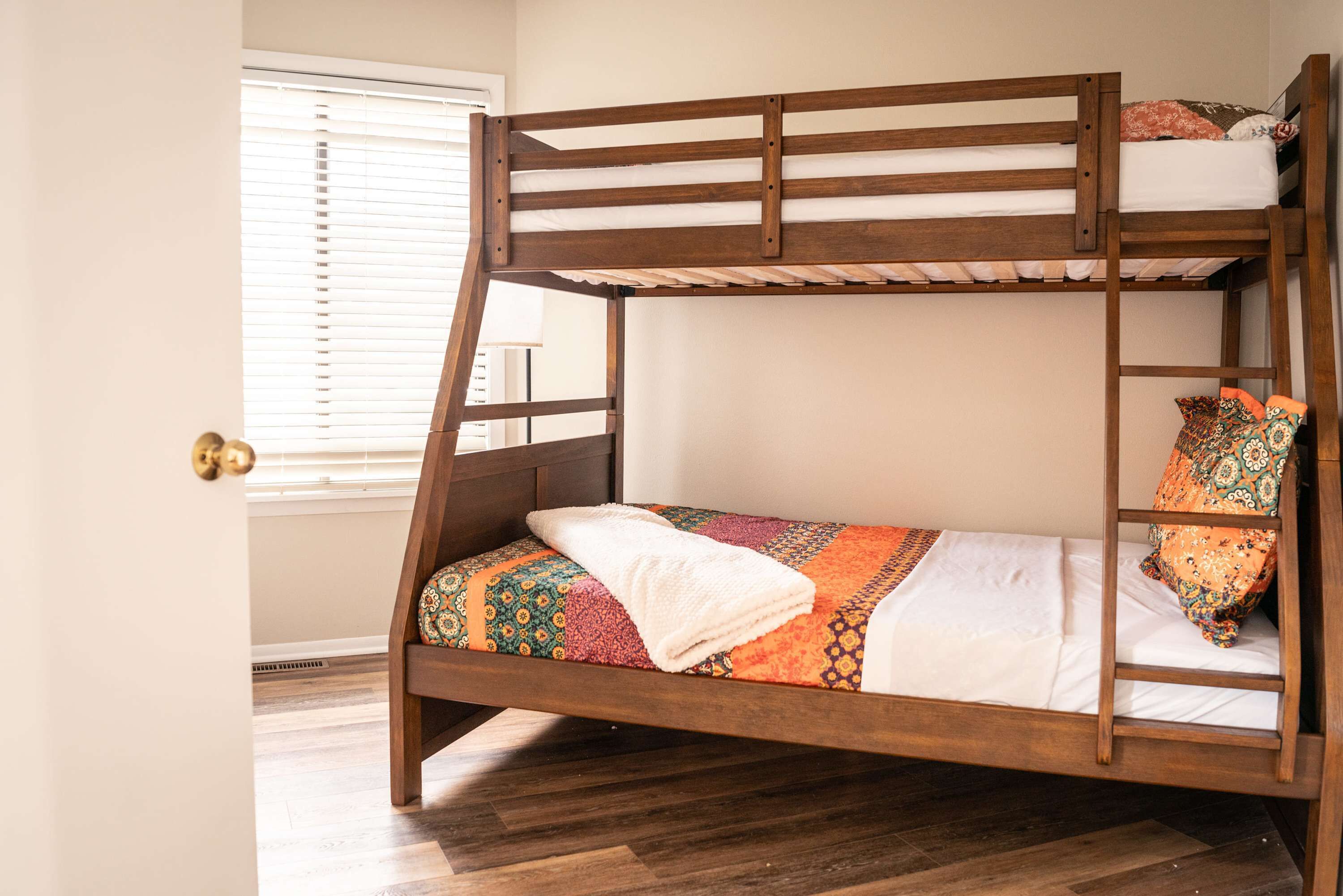 A bunk bed with a ladder, a patterned bedspread, and a window with blinds.