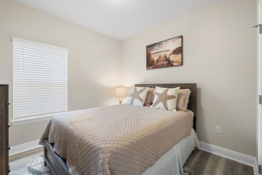 A bedroom with a bed, a window with blinds, a wall-mounted picture, and a nightstand with a lamp.