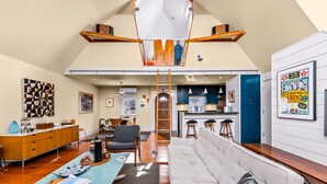 A loft-style living space with a high ceiling, a dining area, kitchen, and a staircase leading to an upper level. - *NEW* Historic Ptown Cottage - Dogs Welcome, Deck, Parking, Vibrant and Colorful (Provincetown)