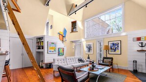 A spacious living area with a staircase, a sofa, a coffee table, and artwork on the walls. - *NEW* Historic Ptown Cottage - Dogs Welcome, Deck, Parking, Vibrant and Colorful (Provincetown)