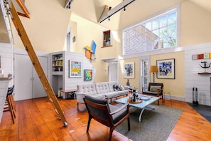 A spacious living area with a staircase, a sofa, a coffee table, and artwork on the walls. - *NEW* Historic Ptown Cottage - Dogs Welcome, Deck, Parking, Vibrant and Colorful (Provincetown)