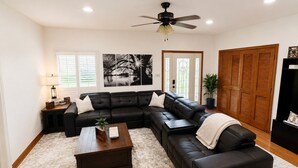 A living room with a sectional sofa, a coffee table, a ceiling fan, and a large window with blinds. - The Bass Shack (Alba)