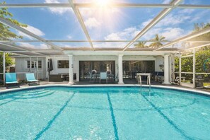 A clear blue pool with a sunlit sky, white columns, and a covered seating area with chairs and tables.