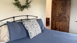 A bedroom with a bed, a wooden door, a wall-mounted wreath, and a ceiling light fixture. - Country Cottage - New Construction On 20 acres (Miles City)