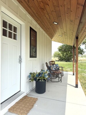 A covered porch with a wooden ceiling, a white door, and a potted plant. - Country Cottage - New Construction On 20 acres (Miles City)