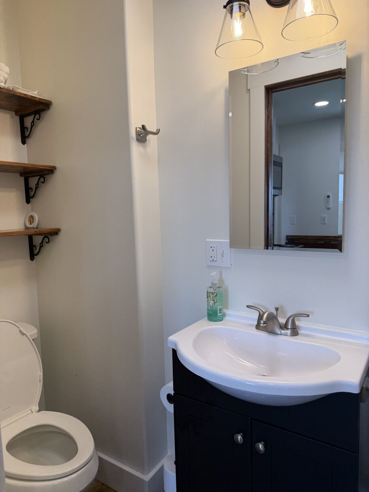 A bathroom with a white sink, black cabinet, and a mirror above it. There are shelves on the wall and a toilet visible.