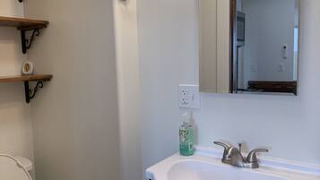 A bathroom with a white sink, black cabinet, and a mirror above it. There are shelves on the wall and a toilet visible.