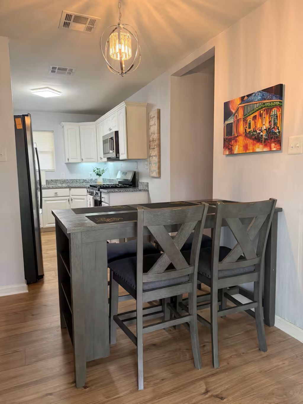 A modern kitchen with white cabinets, a dining area with a wooden table and chairs, a hanging light fixture, and a painting on the wall.