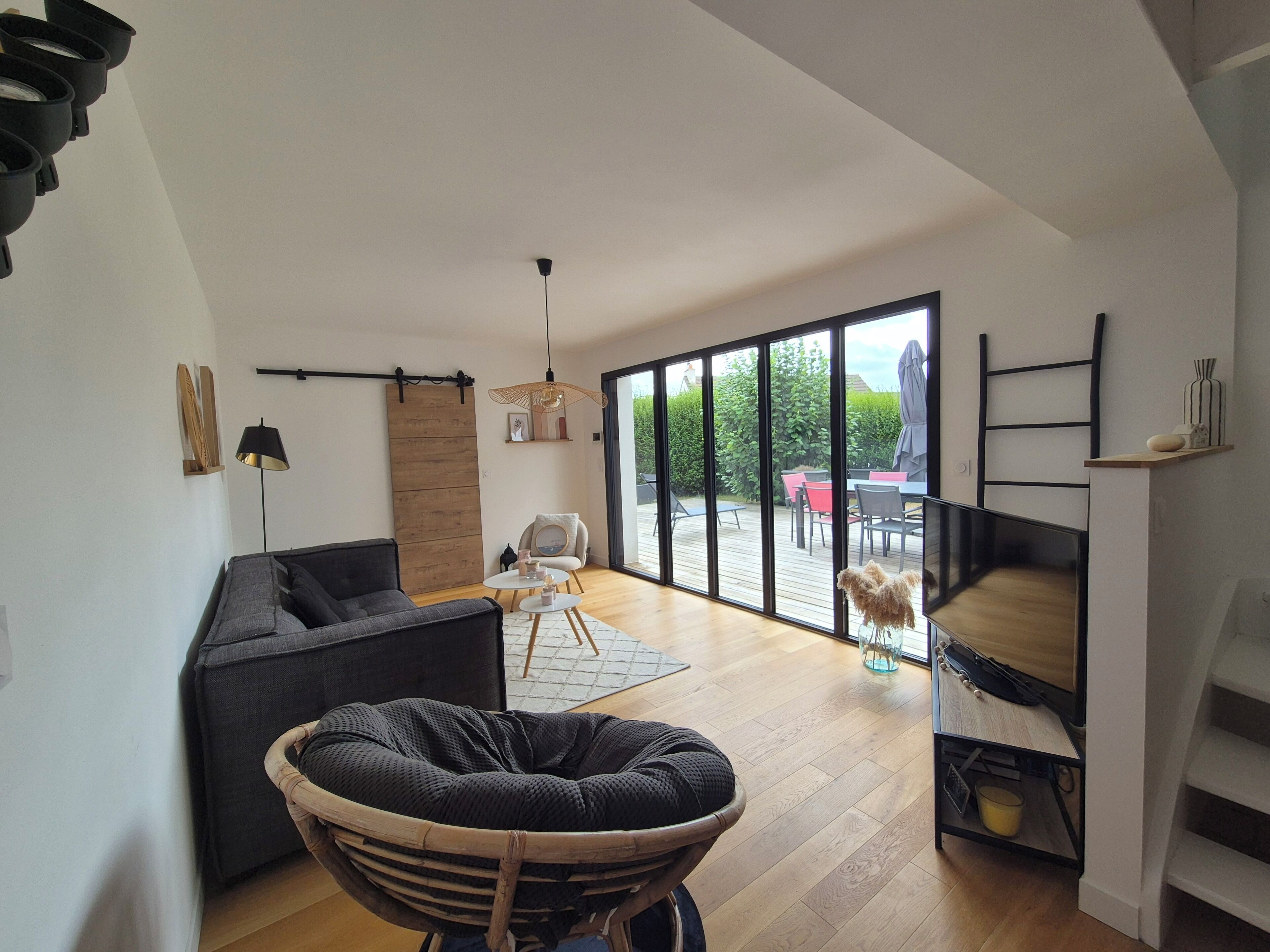 Ein modernes Wohnzimmer mit einer Couch, einem Sessel, einem Essbereich mit Tisch und Stühlen sowie einer großen Glasschiebetür, die auf eine Terrasse im Freien führt.