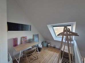 A room with a sloped ceiling, a wooden desk, a wicker chair, a window, and a hanging rack.