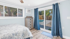 A bedroom with a bed, a dresser, a ceiling fan, and a view of a backyard with a pool. - Serene 1-bedroom  in St. Pete Beach - UNIT B (St. Pete Beach)