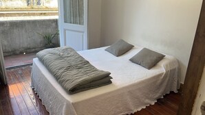 Un lit recouvert d’une courtepointe blanche, avec deux oreillers, une plante en pot et une vue sur un immeuble par la fenêtre.
