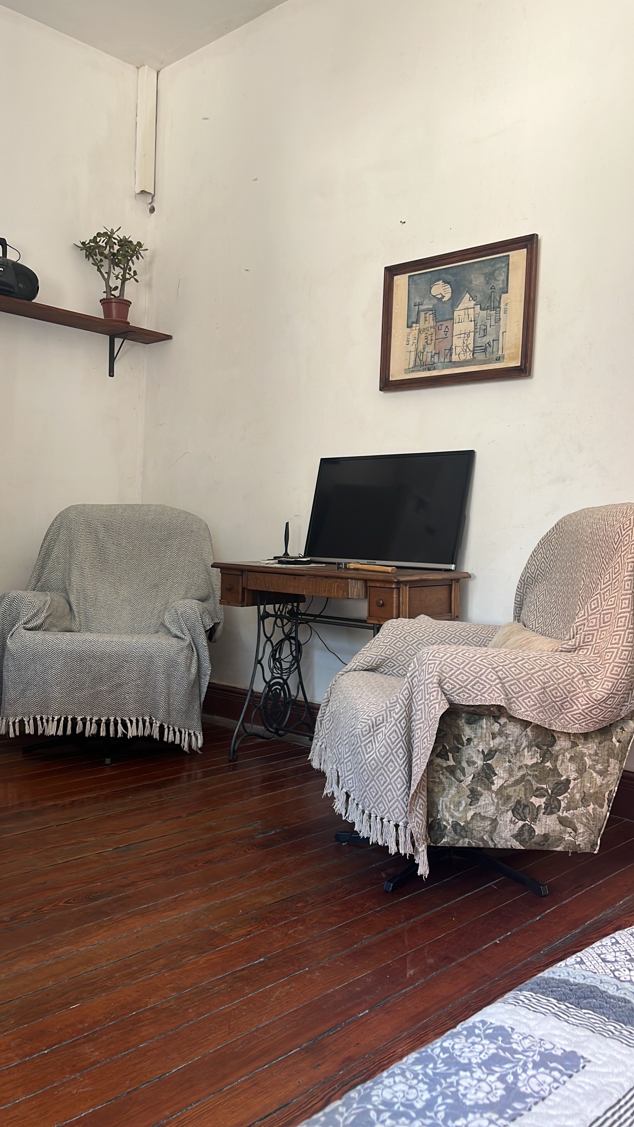 Una habitación con dos sillones, un escritorio de madera con una computadora portátil, un cuadro enmarcado de un paisaje urbano y una planta en maceta.