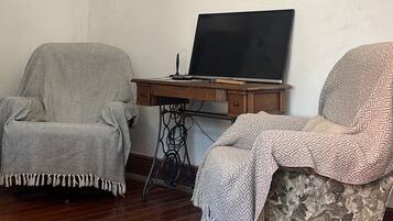 Una habitación con dos sillones, un escritorio de madera con una computadora portátil, un cuadro enmarcado de un paisaje urbano y una planta en maceta.