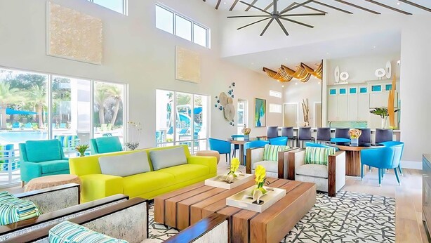 A spacious living area with a bright yellow sofa, wooden coffee table, and blue chairs. A kitchen area with modern appliances and blue chairs is visible in the background. - Upstay Sleeps 26 w Private Pool, Hot Tub, Games (Kissimmee)