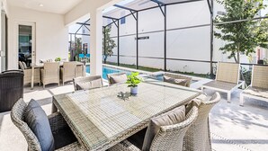 A covered outdoor dining area with wicker furniture and a glass table, overlooking a swimming pool and a white building. - Upstay Sleeps 26 w Private Pool, Hot Tub, Games (Kissimmee)