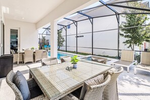 Ein überdachter Essbereich im Freien mit Korbmöbeln und einem Glastisch mit Blick auf einen Pool und ein weißes Gebäude.