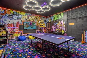 Ein Raum mit einem Pongtisch, einem farbenfrohen Teppich mit Sternenmuster, Graffiti-Wänden und einem Fernsehbildschirm.