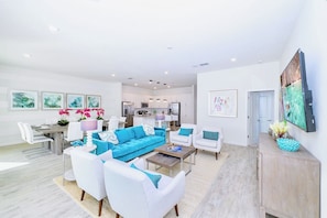 A modern living room with a blue sofa, white armchairs, and a coffee table. There is a dining area with a white table and chairs, and a kitchen in the background. - Upstay Mansion w Cinema, Games, Pool- Sleeps 24! (Kissimmee)