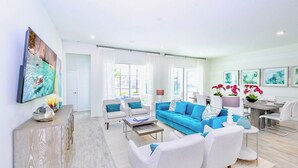 A spacious living room with a blue sofa, white armchairs, and a coffee table. There is a dining area with a table and chairs, and a kitchen in the background. - Upstay Mansion w Cinema, Games, Pool- Sleeps 24! (Kissimmee)