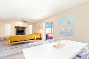 A modern living room with a yellow sofa, a fireplace, a ceiling fan, and a dining area with a white table.