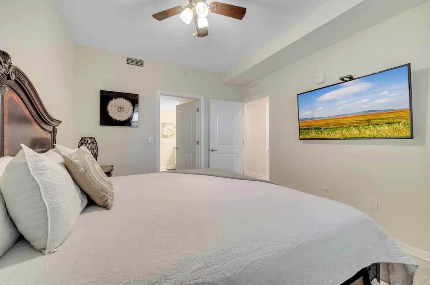 Un dormitorio con una cama grande, un ventilador de techo, un televisor instalado en la pared y una puerta que da a otra habitación.