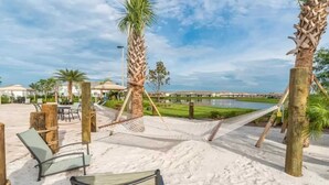 Una hamaca entre palmeras sobre una playa arenosa, con una piscina y sillas de fondo.
