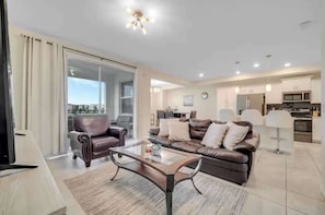 A modern living room with a sofa, armchair, and coffee table, a kitchen area with appliances, and a sliding glass door to an outdoor balcony. - Storey Lake Resort Condo w Patio5 Miles to Disney (Kissimmee)
