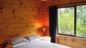 A wooden cabin room with a bed, a nightstand, a lamp, and a window with a view of greenery. - Puka Lodge (Front dwelling) 2 mins walk to beautiful Pukawa Bay Beach Lake Taupo (Pukawa)