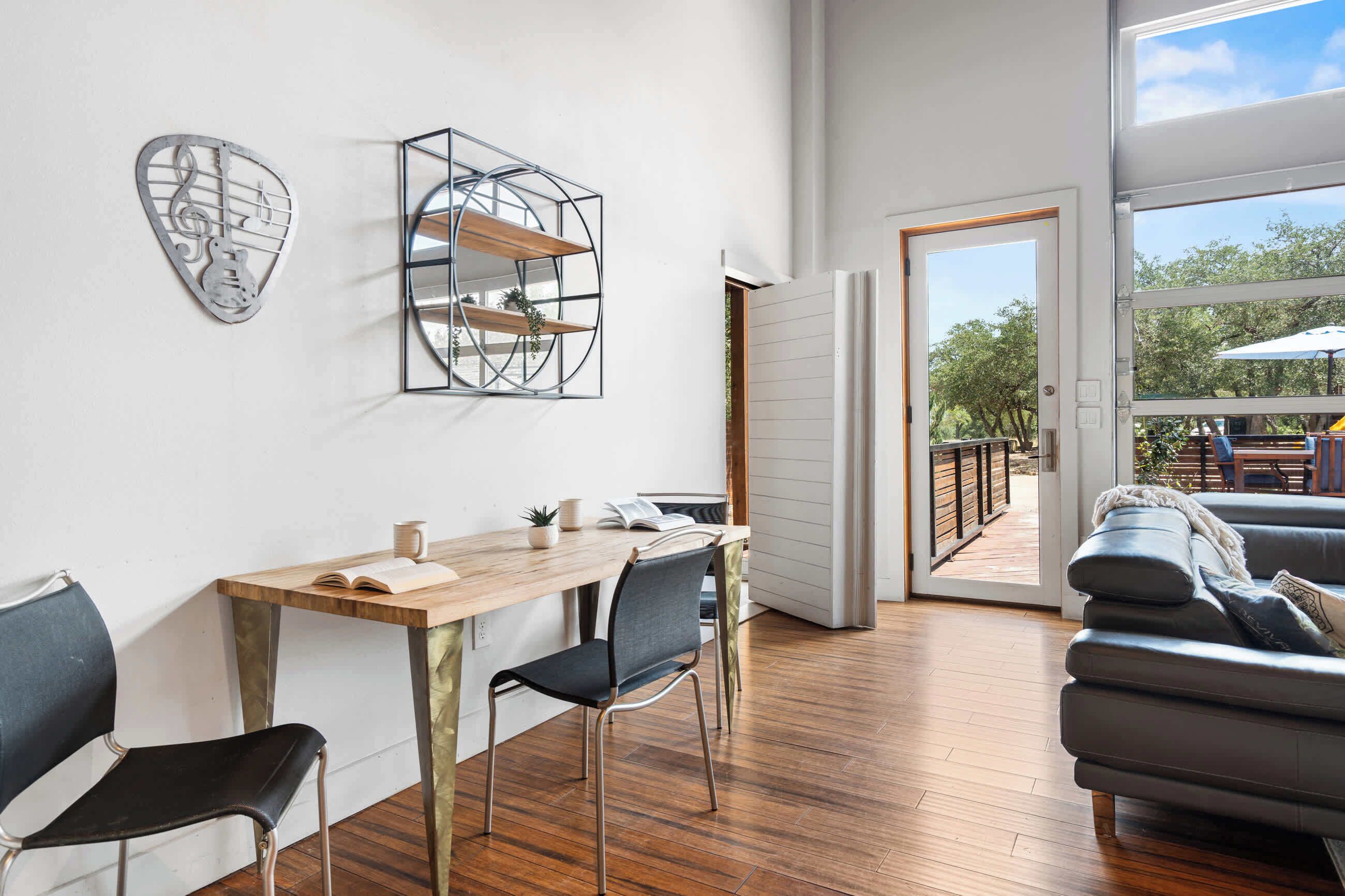 A modern living room with a wooden dining table, two chairs, a sofa, and a large window offering a view of the outdoors.