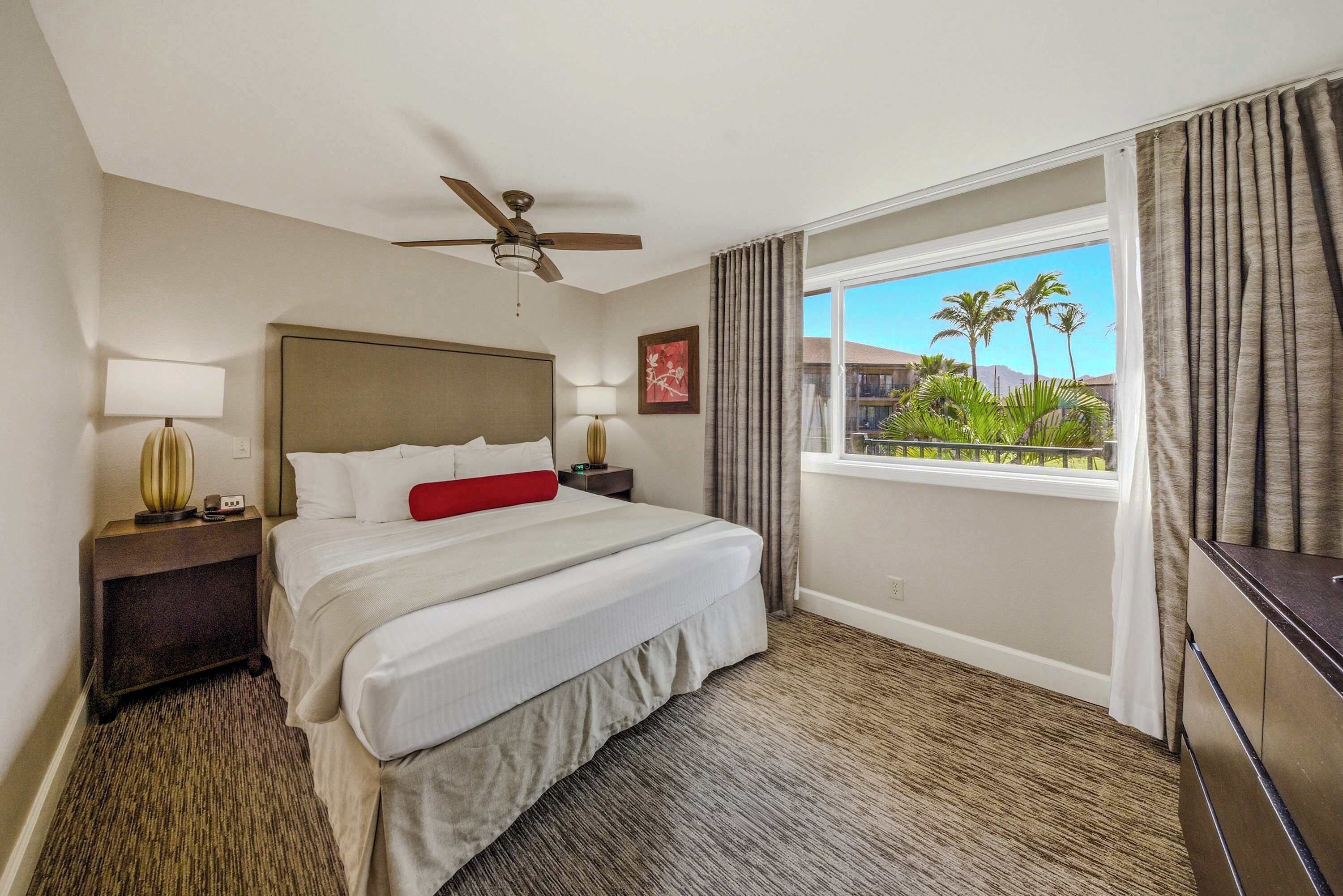 Una habitación de hotel con una cama, ventilador de techo, ventana con cortinas y vista a palmeras.