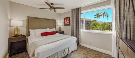 Una habitación de hotel con una cama, ventilador de techo, ventana con cortinas y vista a palmeras.