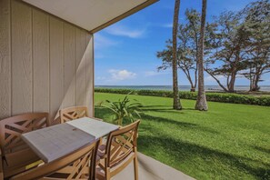 A wooden table and chairs on a balcony with a view of a well-maintained lawn and tall trees. - Oceanfront Escape | Pono Kai Resort | A103 (Kapaʻa)