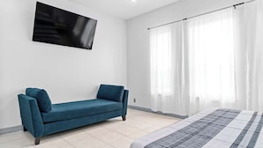 Ein modernes Schlafzimmer mit einem blauen Sofa, einem Bett mit gestreifter Bettdecke, einem großen Fenster mit durchscheinenden Vorhängen und einem am Wand montierten Flachbildfernseher.