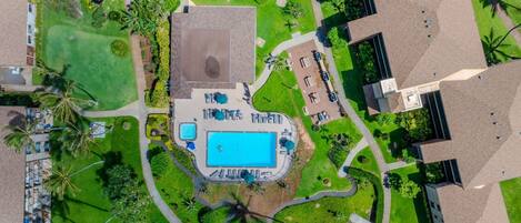 An aerial view of a resort with a central swimming pool, surrounded by pathways, palm trees, and modern buildings.