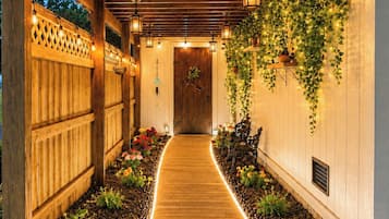 A wooden pathway leading to a door, flanked by a wooden fence and a wall with climbing plants and string lights.