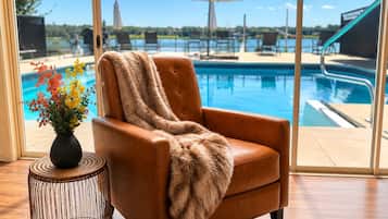 A tan leather armchair with a fur throw, a wicker side table with a vase of flowers, and a view of a swimming pool with lounge chairs and umbrellas.