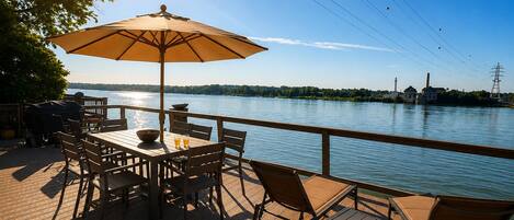 Een terras met een tafel, stoelen, een grote parasol en uitzicht op een waterpartij met gebouwen op de achtergrond.