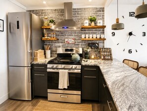 A modern kitchen with a stainless steel refrigerator, a gas stove, and a built-in oven. There are wooden shelves with kitchen items and a clock on the wall. - Romantic Waterfront Views BBQ HotTub Fireplaces Private Pool Pets Luxe Amenities (Jeffersonville)