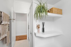 Um corredor com uma porta branca, uma planta pendente, uma prateleira com uma cesta de vime e um vaso com flores secas.