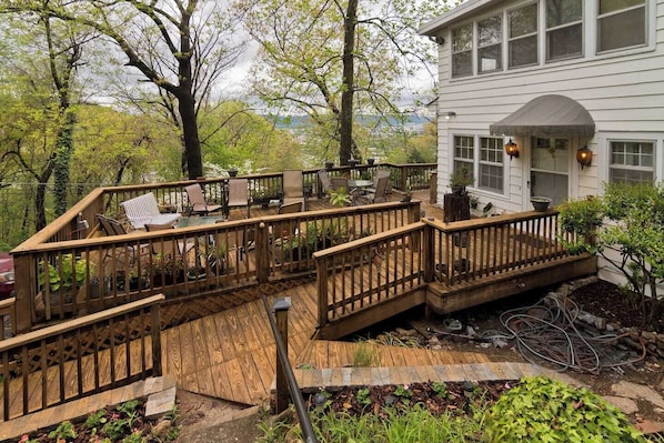 A wooden deck with chairs and tables, a white building with a porch, and a view of trees and hills.
