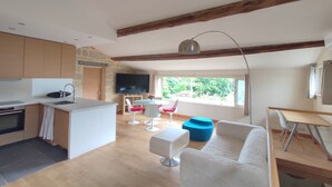 Une cuisine moderne avec un mur en pierre, des poutres en bois, un grand téléviseur et un coin repas avec des chaises blanches et bleues.