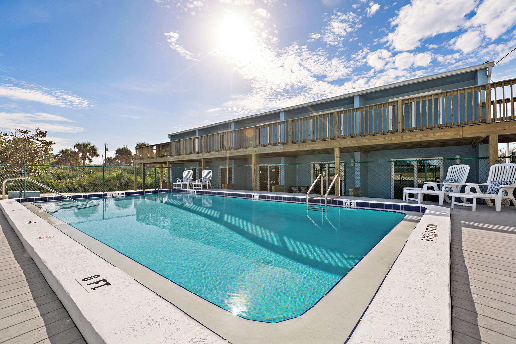 Una piscina rectangular amb una terrassa de fusta al voltant, una terrassa de fusta amb barana i un edifici amb un balcó al fons.