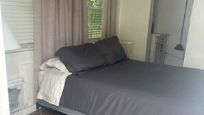 A bedroom with a bed, a window with curtains, a radiator, a desk, and a lamp. - Small studio (West Palm Beach)
