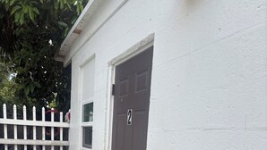 A white building with a brown door marked with the number 2, a white picket fence, and a concrete ground with scattered leaves. - Small studio (West Palm Beach)