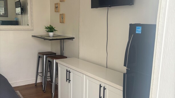 A compact kitchen with a white countertop, a black refrigerator, a mounted TV, and a small table with a potted plant. - Small studio (West Palm Beach)