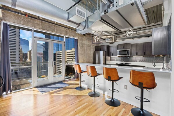 A modern kitchen with a bar area, large windows, and a city view. - Clocktower Views | Sky Lofts 1BR | 30+ Stay (Denver)